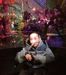 an Asian American disabled woman in a wheelchair in front of a pool of water with tropical water plants in the background lit with purple & magenta lights inside a greenhouse. She is wearing a puffy gray jacket and a mask over her nose attached to a tube.