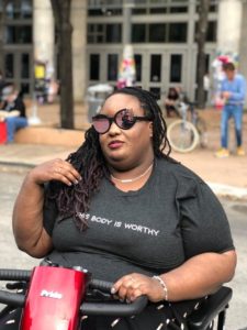A Black woman sits on a mobility scooter that says "Pride" on the front. She's outside on a sunny day, and another person in a wheelchair is reflected in her sunglasses. She lusciously sweeps away a few long dreadlocks to reveal words on her gray t-shirt, "This body is worthy."