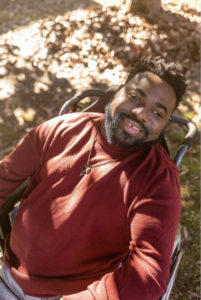 A fall day. AJ, a Black man with a beard and his black hair cut into low temp fade twist-out afro, sits in a manual wheelchair smiling in the dappled sunlight. He wears a burgundy crewneck sweater, plaid dress pants, and a long length leather necklace with an arrow, bulletproof symbol, and jade stone medallion.