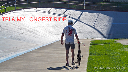 A man in bike clothes, walks down the long track with his fixed gear track bike at the Alpenrose Velodrome on a bright, sunny day. His shirt has a custom-designed logo of a bike cog wheel inspired by a Yin Yang symbol and "Team Kajamo" in red. Text on the picture: "TBI & My Longest Ride: My Documentary Film."