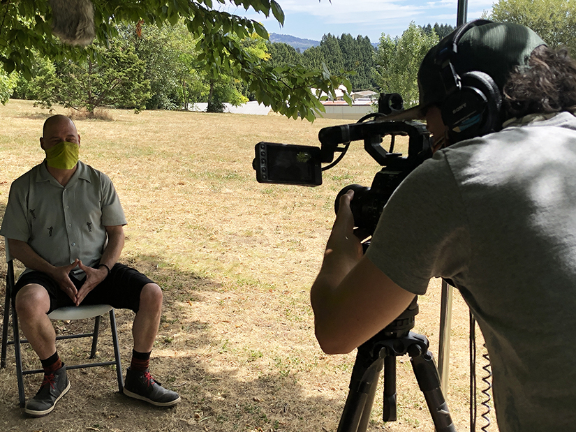 Karl sits in the shade of a tree wearing a fabric face covering. Kai, the videographer, leans into his video camera, focusing the shot.