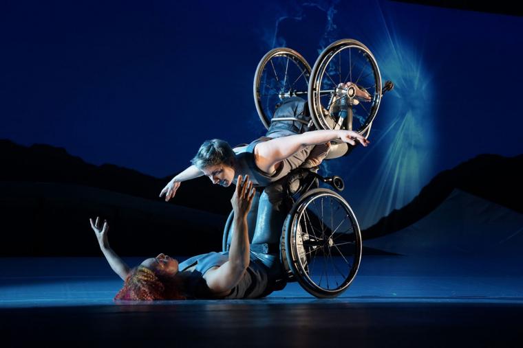 Description courtesy of Kinetic Light: Laurel Lawson, a white woman, is flying in the air with arms spread wide, wheels spinning, and supported by Alice Sheppard. Alice, a light-skinned Black woman, is lifting from the ground brelow. They are making eye contact and smiling. Photo by Jay Newman / BRITT Festival.