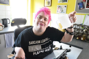 A white woman with neon pink sits in a power wheelchair with a communication device mounted on the front. She's smirking and holding up a white plastic coffee cup with two handles. Her shirt says "Sarcasm loading, please wait," with an online page loading progress bar showing loading is only halfway done.