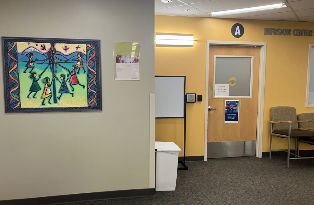 A painting of children dancing around a maypole hangs on the wall by the entrance into a hospital infusion center. Bright fluorescent lights blare above the door to the center.
