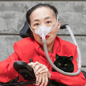 Alice Wong, an Asian American woman with short black hair, sits in a power wheelchair with her hands crossed and draped over the joystick panel. She has a BiPAP mask over her nose with a hose connected to her vent behind her, out of view. Her bold red lip matches a high-shoulder-padded red blouse with a scarf at the neck. A black cat with fierce yellow eyes is composited onto her lap, tucked under her left arm. He's positioned perfectly to bite her BiPAP tube, but he's not. He's leaning contentedly into the crook of her elbow.